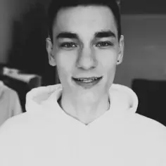 Uśmiechnięty chłopak, chati youtuber, w białej bluzie z kapturem, w tle widać inne osoby.
