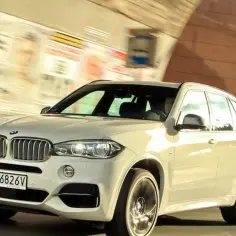 Białe BMW X5 M50d pędzi ulicą pod wiaduktem.
