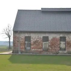 Stary dom z obory, z ceglaną ścianą i szarym dachem, otoczony zieloną trawą i drzewami.