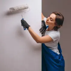 Kobieta w niebieskim ogrodniczkach nakłada wałkiem grunt pod styropian na ścianę.