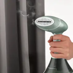 Dłoń trzyma parownicę Russell Hobbs, z której wydobywa się para, zbliżając ją do szarych zasłon.