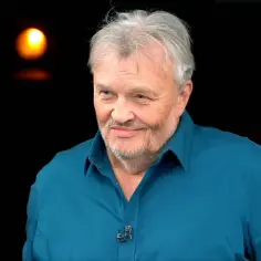 Krzysztof Cugowski, w niebieskiej koszuli, z siwą brodą i włosami, uśmiecha się. W tle migoczą światła, przypominając o jego wspaniałych utworach.