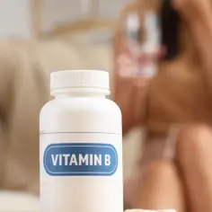 Biała butelka z etykietą "VITAMIN B" stoi na stole. W tle kobieta pije wodę. Tiamina, składnik witaminy B, wspiera metabolizm.