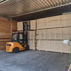 Pomarańczowy wózek widłowy przygotowuje stosy desek do suszenia drewna w suszarni.