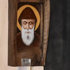 Obraz przedstawia portret św. Charbela w brązowym habicie i kapturze, z aureolą. Obok widoczna jest drewniana rama, sugerująca, że jest to część relikwiarza lub ikonostasu, być może związana z przepowiedniami św. Charbela.