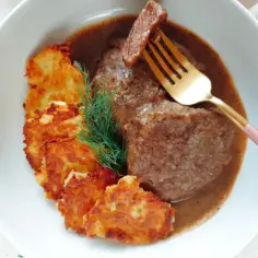 Bitki wołowe w sosie z plackami ziemniaczanymi i gałązką koperku, na talerzu.