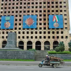 Zniszczony budynek w Abchazji z wielkimi banerami z medalami. Na pierwszym planie mężczyzna na skuterze z przyczepą pełną drewna.