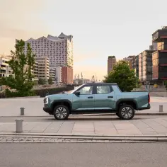 Nowoczesny, turkusowy Maxus pickup stoi na tle portowego miasta o zachodzie słońca.