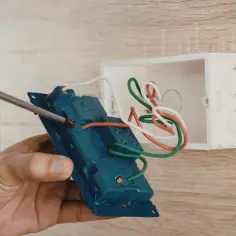 Dłoń przykręca śrubokrętem niebieski element instalacji elektrycznej z przewodami do białej puszki.