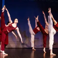 Grupa tancerzy w bordowych i białych strojach wykonuje dynamiczne figury na scenie. Każdy tancerz prezentuje wysokie uniesienie nogi.