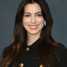 Anne Hathaway w czarnej marynarce z ozdobnymi guzikami, uśmiecha się do aparatu.