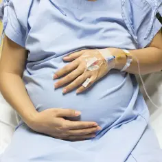 Kobieta w ciąży w szpitalnej koszuli z kroplówką. Dłonie na brzuchu, być może odczuwa objawy odklejenia łożyska.