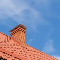 Ceglany komin na czerwonym dachu pod błękitnym niebem z chmurami.
