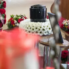 Czarna urna na wieńcu z białych kwiatów, otoczona bukietami. Czy kremacja boli zmarłego? Nie, to proces bezbolesny.