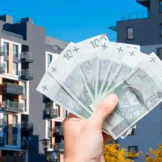 Dłoń trzyma polskie banknoty 100 zł na tle nowoczesnych budynków mieszkalnych. Może to symbolizować podatek od sprzedaży nieruchomości.