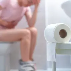 Osoba siedząca na toalecie, trzymająca się za brzuch, z rolką papieru toaletowego w pobliżu. Może to być pomocne przy problemach z brzuszkiem, jak przy stosowaniu meteospasmyl.