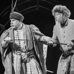 Dwóch mężczyzn w historycznych strojach, jeden z szablą, drugi z fuzją. Ich miny sugerują komizm w zemście, gdy przygotowują się do konfrontacji.