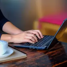 Dłonie piszą na laptopie, obok filiżanka kawy i notatnik. Czas na odliczenie internetu i pracę.