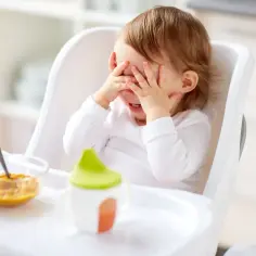 Małe dziecko w krzesełku zasłania oczy rączkami, wyraźnie widać, że **noworodek nie chce jeść**. Obok miseczka z jedzeniem i kubeczek.