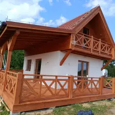 Nowy biały dom z drewnianym gankiem i balkonem, otoczony zielenią.