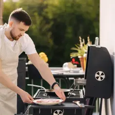 Mężczyzna w fartuchu przygotowuje mięso na grillu, pokazując, że można grillować na balkonie.