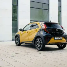 Żółto-czarny Toyota Aygo X Hybrid zaparkowany na placu przed nowoczesnym budynkiem.