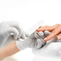 Dłonie w białych rękawiczkach piłują paznokcie, pokazując, jak zdjąć tipsy. Na białym ręczniku leży dłoń z delikatnym manicure.