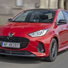Czerwona Mazda 2 Hybrid w ruchu miejskim, z widocznym grillem i reflektorami.