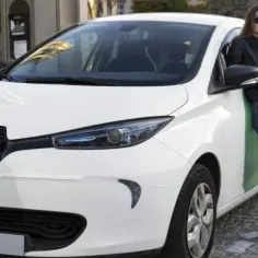 Elegancka kobieta rozmawia przez telefon, opierając się o biały samochód elektryczny do 30 tys. zł, który się ładuje.