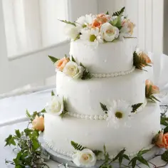 Elegancki tort weselny udekorowany białymi i brzoskwiniowymi różami, margaretkami i zielonymi liśćmi. Inspiracja, jak udekorować tort kwiatami.