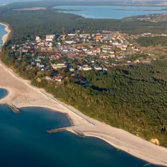 Widok z lotu ptaka na polskie wybrzeże z piaszczystą plażą, kamiennymi falochronami i lasem sosnowym. W oddali widać zabudowania, jezioro i farmę wiatraków.