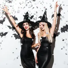 Dwie kobiety w strojach czarownic, z czarnymi kapeluszami i sukienkami, świętują Halloween. Konfetti i nietoperze na ścianie dodają klimatu. Idealny pomysł, jak zrobić strój diabła na imprezę!