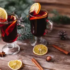 Dwa kubki aromatycznego grzańca z whisky, ozdobione plastrami pomarańczy i laskami cynamonu, na drewnianym stole wśród gałązek świerku.