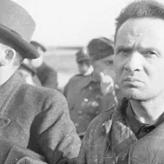 Dwóch mężczyzn, jeden w kapeluszu, drugi z surowym wyrazem twarzy, stoi w tłumie. Być może to komendant i jego podwładni.