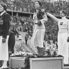 Trzy medalistki na podium, wśród nich triumfatorka z wieńcem laurowym. Scena przypomina pierwsze igrzyska olimpijskie.