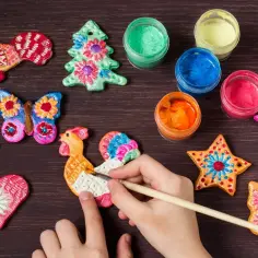 Dziecko maluje koguta z masy solnej, tworząc kolorowe inspiracje. Obok ozdoby choinkowe, motyle i gwiazdki.