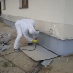 Osoba w kombinezonie maluje cokół budynku metodą natryskową. To pokazuje, jak malować elewację, by uzyskać równomierne krycie.