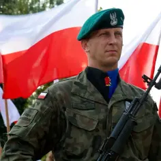 Żołnierz w zielonym beretcie, z karabinem i polskimi flagami w tle. Czy z licencjatem można zostać oficerem? Tak, to ścieżka kariery w wojsku.