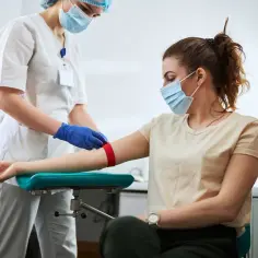 Pielęgniarka w maseczce i rękawiczkach zakłada opaskę uciskową na ramię pacjentki, przygotowując ją do pobrania krwi. Może to być związane z podwyższonym ob.