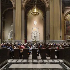 Ludzie siedzą w ławkach w kościele, słuchając kazania. Wnętrze świątyni, z ołtarzem i posągiem Matki Boskiej, przypomina architekturę charakterystyczną dla anglikanizmu.