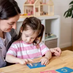 Mama uczy córkę liter, przygotowując ją do przedszkola. Dziecko z uwagą śledzi paluszkiem literkę na niebieskiej karcie.