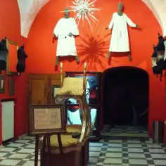 Wnętrze muzeum maszyn erotycznych w Pradze. Czerwone ściany, gorsety na manekinach, wiszące postacie w białych szatach i szachownicowa podłoga.