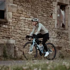 Kobieta na rowerze, testująca nowe marki rowerów, przemierza wiejską drogę obok kamiennego domu.