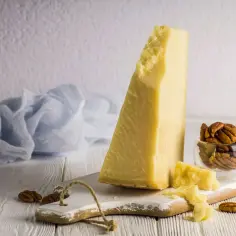Kawałek żółtego sera Grana Padano na desce, obok orzechy pekan i niebieska tkanina.