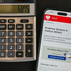Kalkulator i smartfon z aplikacją KSeF. Na ekranie telefonu widoczna jest informacja o Krajowym Systemie e-Faktur, który ułatwia wystawianie i odbiór faktur, także tych bez VAT.