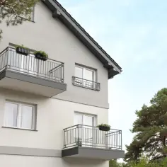 Nowoczesny budynek z balkonami i oknami, otoczony zielenią drzew. Suterena jest niewidoczna, ale całość sprawia wrażenie przytulnego miejsca.