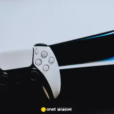 Biały kontroler DualSense leży przed konsolą PlayStation 5. Światło odbija się od jej czarnych i białych elementów, sugerując przyszłość gier.
