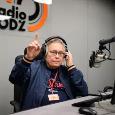 Mężczyzna w słuchawkach i okularach, siedzący przy mikrofonie w studiu radiowym. Wskazuje palcem, jakby coś tłumaczył. Ale numer obsada!
