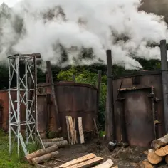 Dym wydobywa się z metalowych pieców do produkcji węgla drzewnego, obok składowisko drewna.
