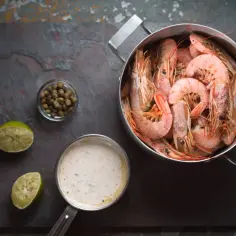 Gotowane krewetki w garnku, sos, kapary i połówki limonki na ciemnym tle.